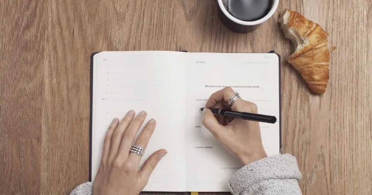 Mains rédigeant des notes dans un carnet de planification, à côté d'un café et d'un croissant