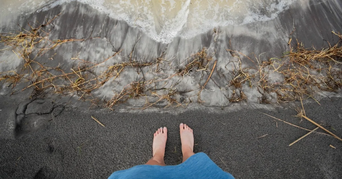 Pieds nus sur le sable gris d'une plage, au bord de l'eau