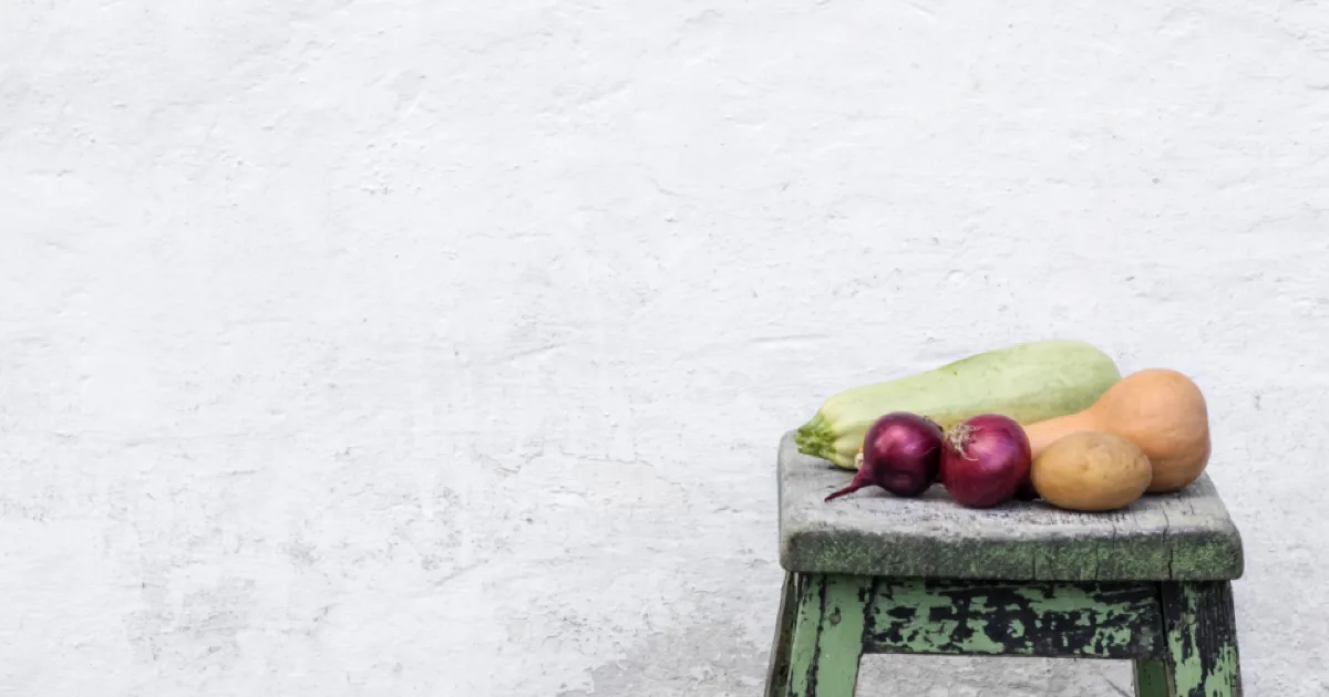 Légumes (courgette, oignons rouges, pomme de terre, courge) posés sur un tabouret ancien devant un mur blanc